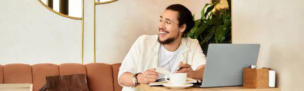 Smiling young man with a beard focuses on his work while sipping coffee in a vibrant cafe, banner — Stock Photo