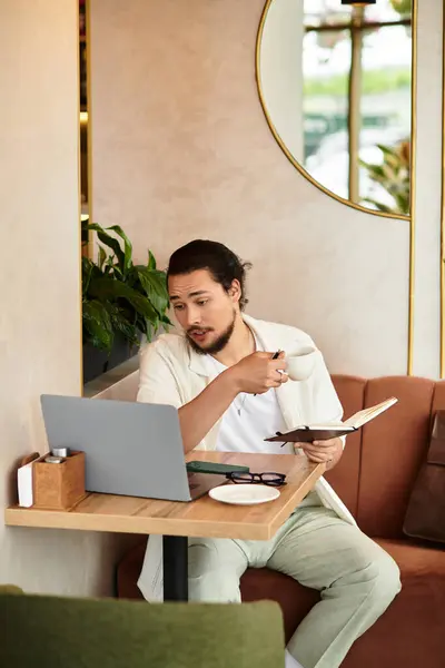 A young man with a beard sits in a cozy cafe, sipping coffee and working on his laptop. — Stock Photo