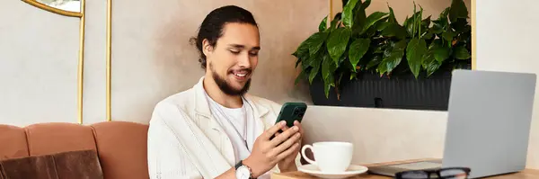A young man with a beard is focused on his smartphone while sipping coffee in a cafe, banner — стоковое фото