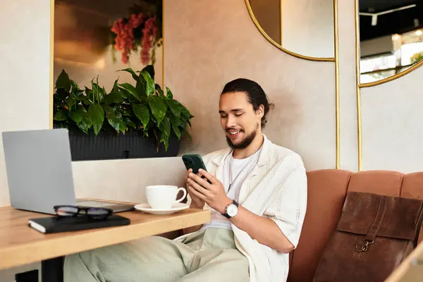 Young man with a beard engages with his phone and laptop in a delightful cafe atmosphere. — Stock Photo