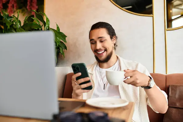 Bearded young man sits in a cafe, smiling as he works on his phone and enjoys a warm drink. — Stock Photo