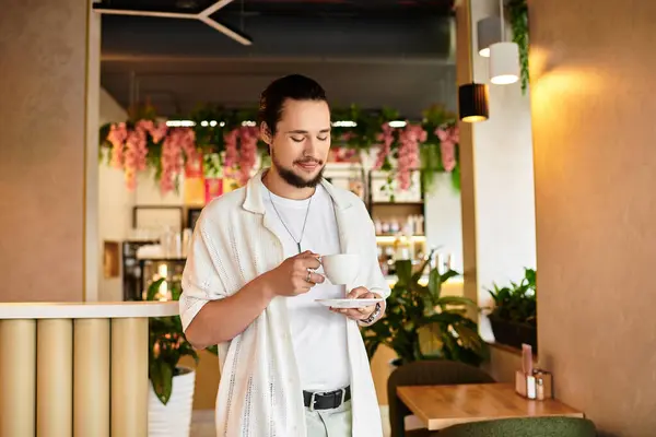 A young man with a beard sips coffee in a cafe surrounded by greenery. — Stock Photo
