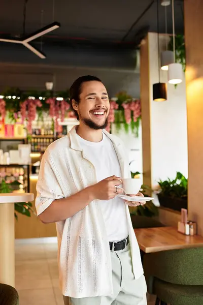 A young man with a beard smiles while holding a cup in a lively cafe. — Stock Photo