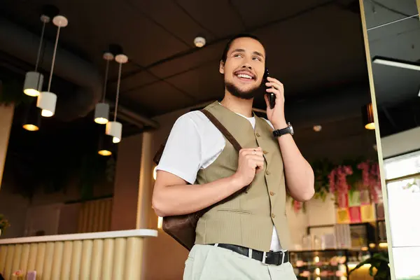 A young man with a beard smiles while using his phone and carrying a bag in a vibrant cafe. — Stock Photo