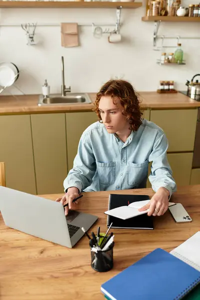 Young man with curly hair sits at a wooden table, engaged in work on his laptop in a modern kitchen. — Stock Photo