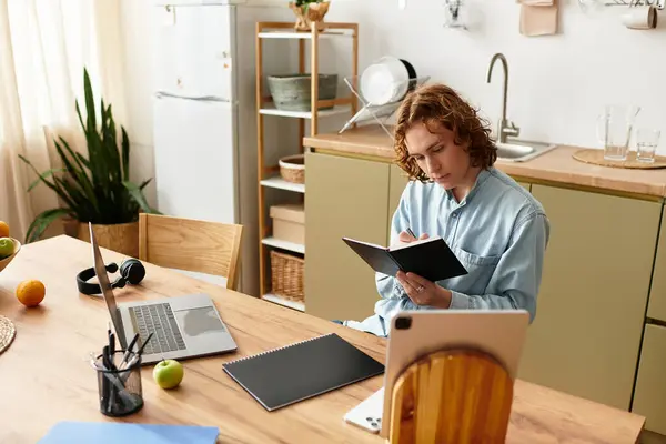 A stylish young man sits at a cozy kitchen table, writing thoughtfully in a notebook. — Stock Photo