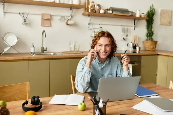 A cheerful young man engages in a phone call while working on his laptop at home during the day. — Stock Photo