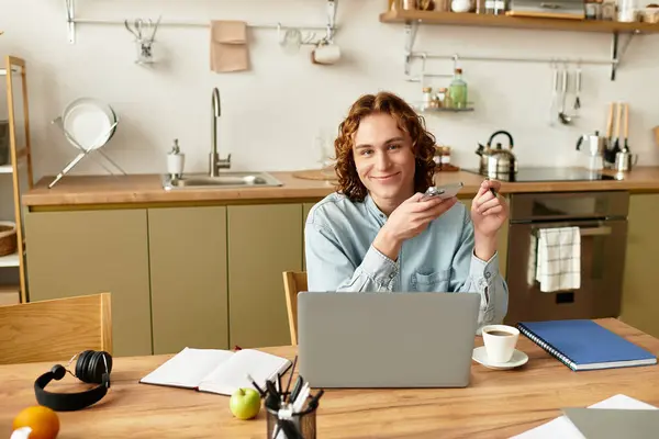 Young man with curly hair is seated at a kitchen table, engaged with his laptop and smiling. — Stock Photo