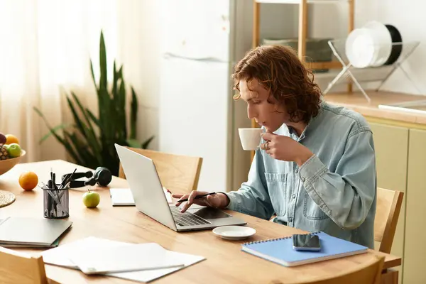 A young, handsome man is seated at a wooden table, focused on his laptop while sipping coffee. — Stock Photo