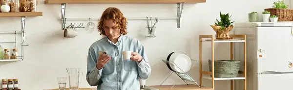 Handsome young man stands in a cozy kitchen, sipping coffee and checking his phone. — Stock Photo