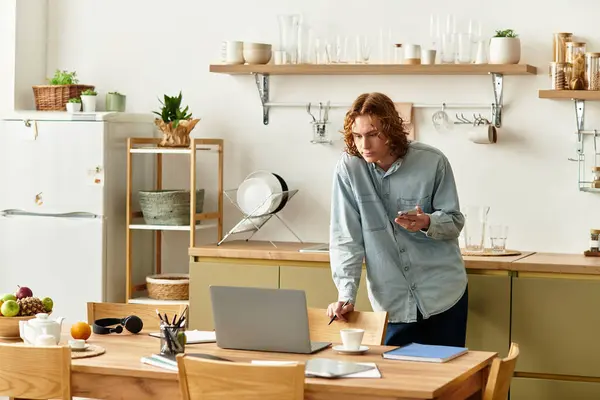 A handsome young man stands thoughtfully at a kitchen table, focusing on tasks with a laptop nearby — Stock Photo
