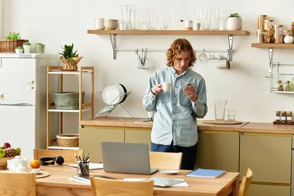 Young man stands in a stylish kitchen, sipping coffee while looking at his phone. — Stock Photo