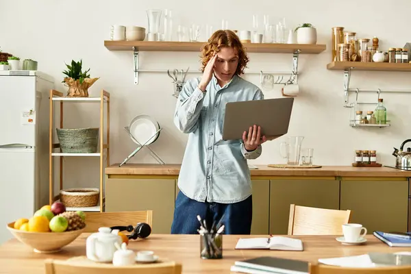 Handsome young man stands in a modern kitchen, absorbed in his laptop as he thinks. — Stock Photo