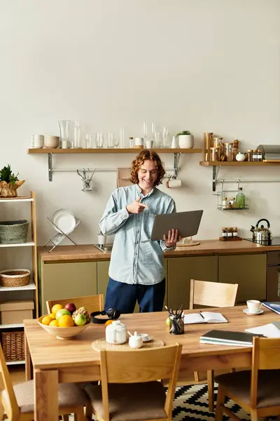 Handsome young man stands in a stylish kitchen, focused on his laptop while smiling. — Stock Photo