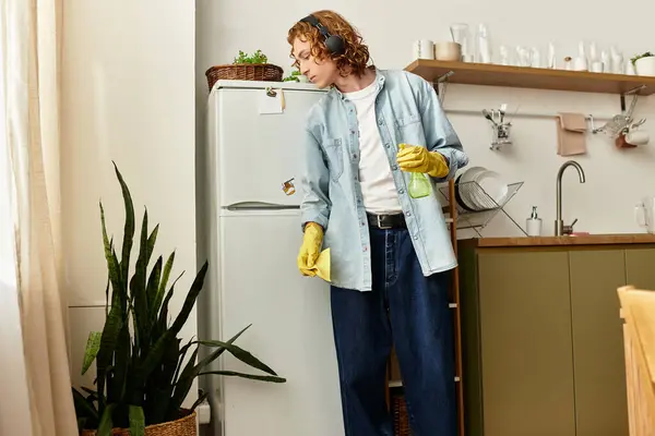 Un joven con el pelo rizado lleva guantes de limpieza, ordenando la cocina en medio de la vegetación. - foto de stock