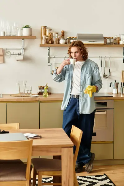 A handsome young man stands in a well lit kitchen, sipping from a cup and relaxing. — Stock Photo