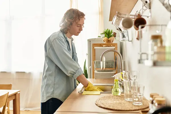 Un joven con el pelo rizado lava platos en una cocina caliente llena de luz natural. - foto de stock