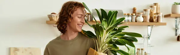 Smiling young man holds a large green plant while enjoying his peaceful home atmosphere. — Stock Photo
