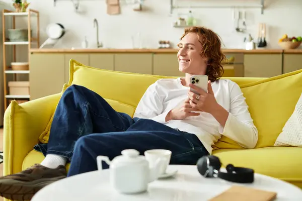 A handsome young man lounges on a bright yellow couch, engaged with his smartphone while smiling. — Stock Photo