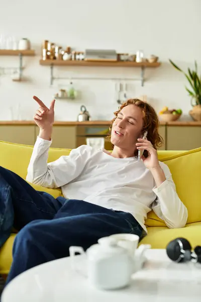 A handsome young man lounges comfortably on a bright yellow sofa, happily chatting on the phone. — Stock Photo