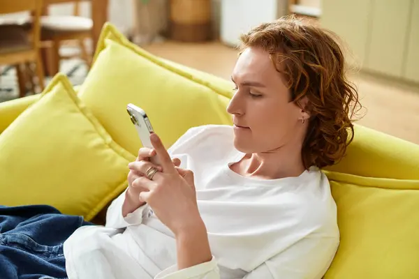 A young man relaxes on a bright yellow couch, absorbed in his smartphone. — Stock Photo