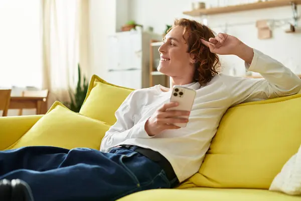 A young man relaxes on a cozy couch, smiling as he uses his phone. — Stock Photo