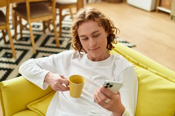 A handsome young man relaxes on a yellow sofa, sipping from a cup and using his smartphone. — Stock Photo