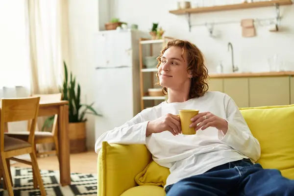 A handsome young man sits on a bright couch, savoring a warm drink and enjoying moments at home. — Stock Photo