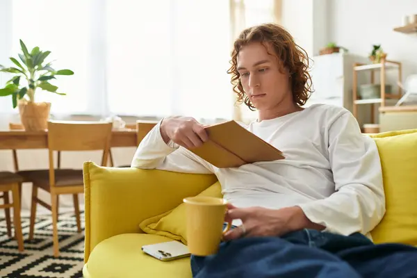A handsome young man sits comfortably at home on a yellow couch, enjoying a book and a warm drink. — Stock Photo