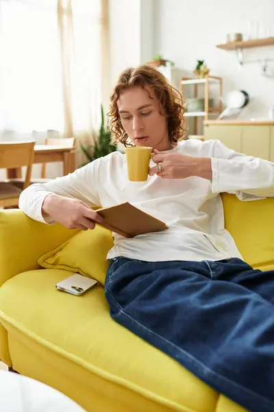 Handsome young man lounges comfortably on a vibrant couch, sipping a warm beverage while reading. — Stock Photo