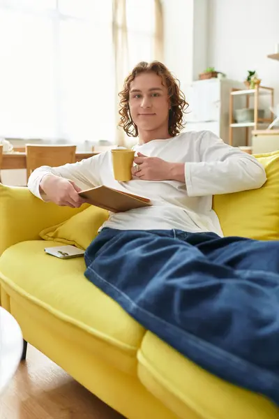 Young man lounges on a bright yellow couch, sipping tea while reading a book in his home. — Stock Photo