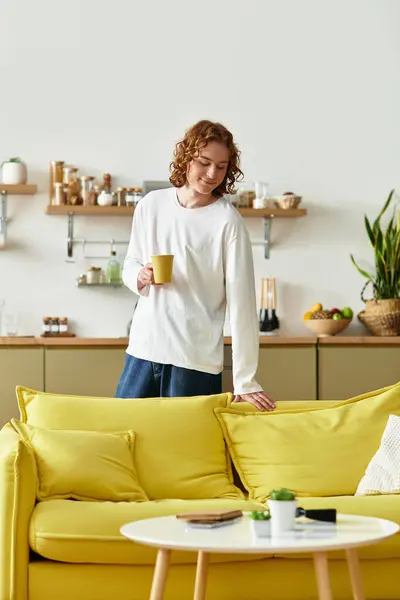 Handsome young man stands by a cheerful yellow sofa, savoring a drink in his inviting living space. — Stock Photo
