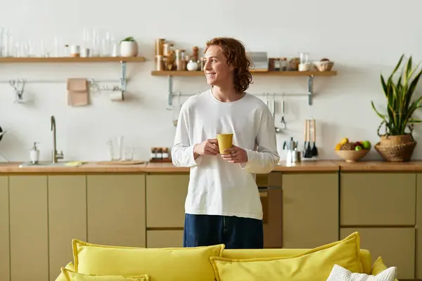 Young man stands in his cozy kitchen, savoring a comforting drink while smiling softly. — Stock Photo