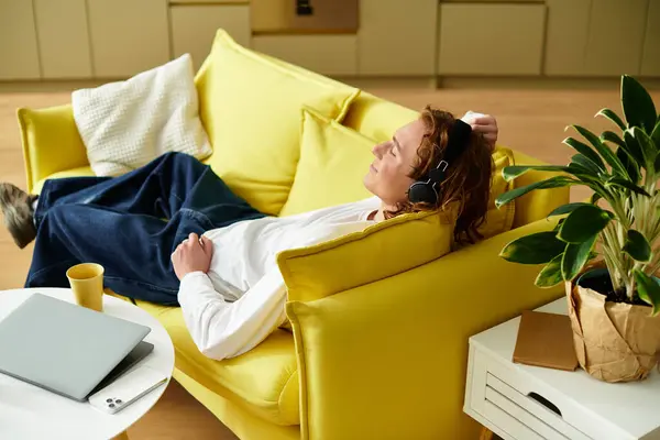 Young man with curly hair listens to music while lounging comfortably on a bright yellow couch — Stock Photo