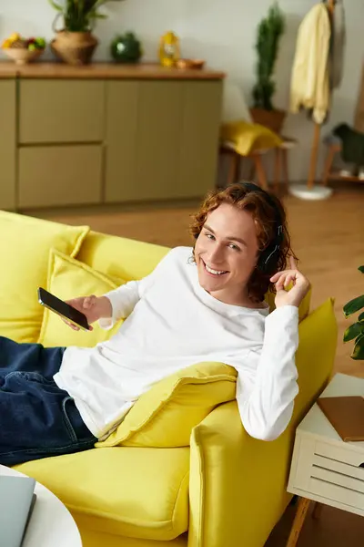 A handsome young man lounges comfortably on a vibrant yellow couch, enjoying his smartphone. — Stock Photo