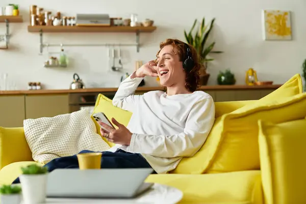 Handsome young man sits comfortably on a bright yellow couch, listening to music and smiling. — Stock Photo