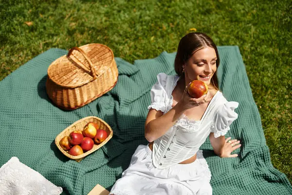A beautiful woman relaxes on a picnic blanket, savoring an apple amidst a lush green park. — Stock Photo