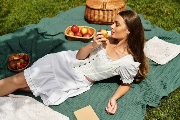 A beautiful young woman relaxes on a picnic blanket, savoring an apple under the warm sun. — Stock Photo