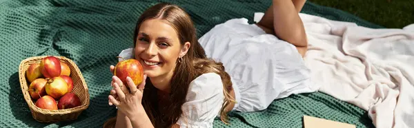 Young woman smiles while lounging on a blanket surrounded by fresh apples during a sunny picnic. — Stock Photo