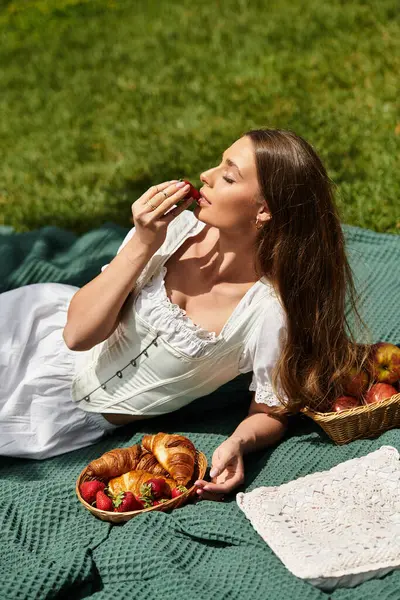 A beautiful young woman indulges in strawberries and croissants while relaxing on a picnic blanket. — Stock Photo