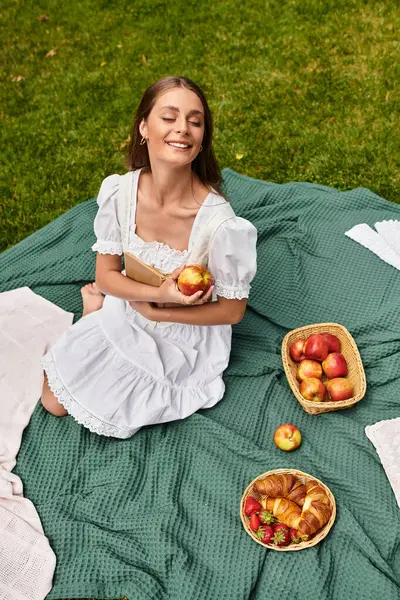 A beautiful young woman sits on a picnic blanket, savoring an apple surrounded by snacks. — Stock Photo
