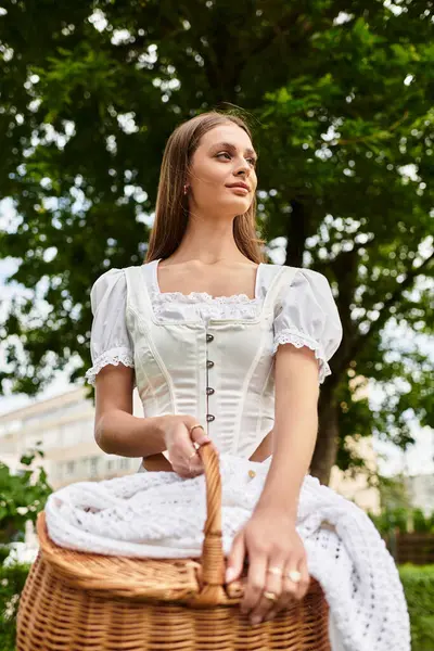A young woman stands confidently in a park, holding a picnic basket amid the greenery. — Stock Photo