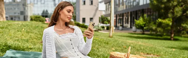 A beautiful young woman sits on a blanket at a park, using her smartphone and enjoying a picnic. — Stock Photo
