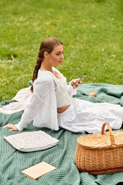 A young woman types on her phone while sitting on a picnic blanket in the park. — Stock Photo
