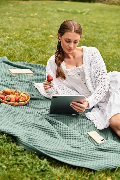Relajarse en un parque mientras está ocupado con una tableta y saborear fresas frescas en una manta de picnic. - foto de stock
