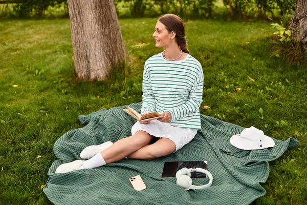Una joven se relaja en una manta de picnic, leyendo un libro en un parque soleado rodeado de vegetación. - foto de stock