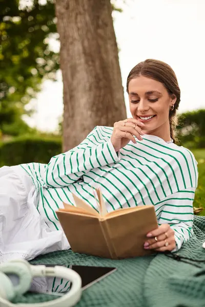 A young beautiful woman enjoys a sunny day, relaxing on a blanket while reading a book in the park. — Stock Photo