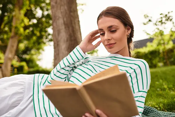 A beautiful woman relaxes on the grass, immersed in her book at a sunny outdoor park. — Stock Photo