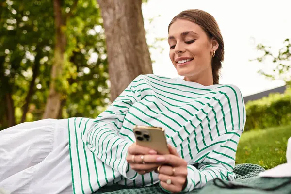 Delightful young woman relaxes on a picnic blanket, engaged with her smartphone under the trees. — Stock Photo