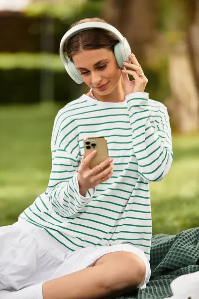 A young woman relaxes on a picnic blanket, enjoying music and the outdoors in a sunny park. — Stock Photo